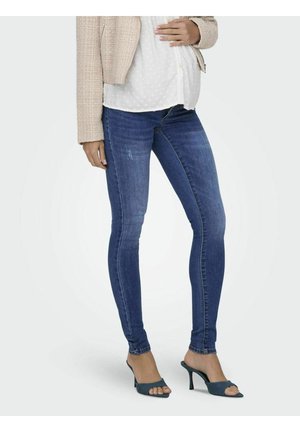 Jeans ajustados de mezclilla azul con un corte clásico, que presentan un ligero desgaste y una cintura de altura media. Combinados con una blusa ligera de lunares y sandalias de tacón.