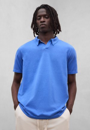 Ung mand med dreadlocks iført en knaldblå kortærmet skjorte med krave og beige bukser med hænderne i lommerne, neutral baggrund.