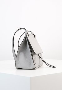 Šedá kožená kabelka trojúhelníkového tvaru, texturovaný povrch, plochý klopový uzávěr a tenký nastavovací popruh, ležící na bílém soklu.