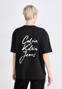 Svart oversized t-skjorte laget av bomull, med en stor hvit håndskrevet logo "Calvin Klein Jeans" på baksiden. Kort ermer, rund hals.