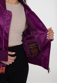 Paarse bomberjack met een satijnen afwerking, gedetailleerd binnenlabel en een knoopzak. Heeft ribbelmanchetten en een ritssluiting.