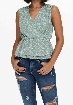 Woman wearing a sleeveless green floral peplum top and ripped dark blue jeans with hands in pockets against a white background.