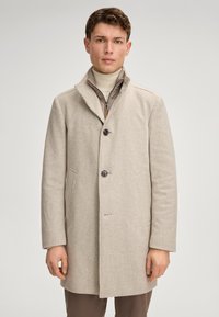 Beige geblokte jas met een notch-kraag, drie knopen, twee zijzakken en een gestructureerde afwerking. Gedragen over een lichte coltrui.