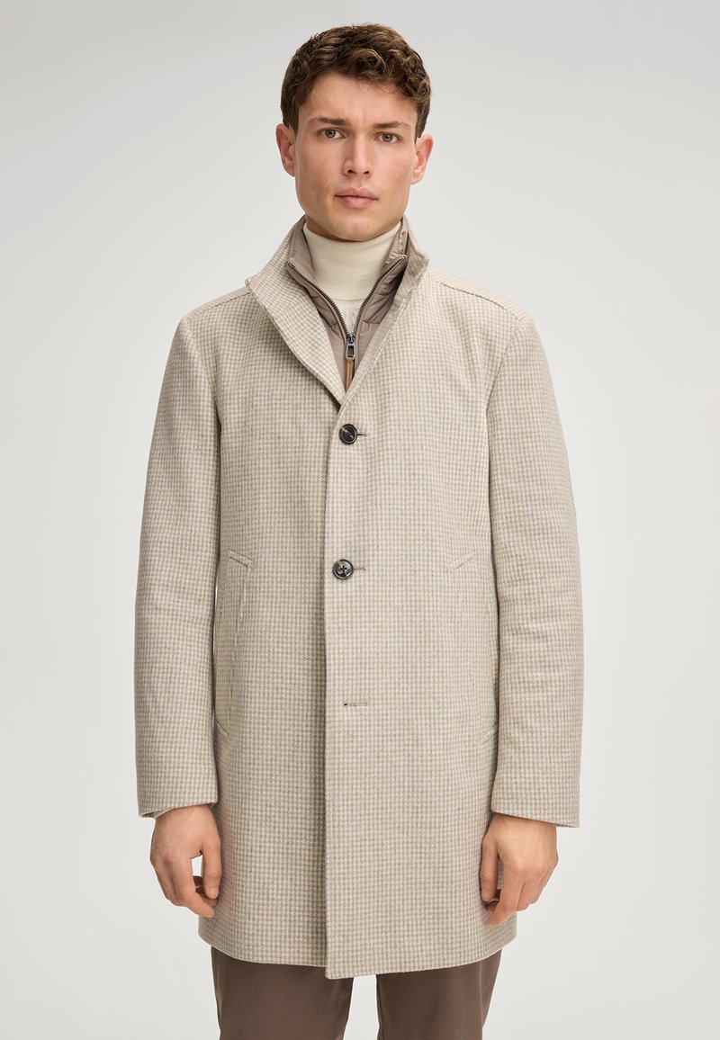 Beige geblokte jas met een notch-kraag, drie knopen, twee zijzakken en een gestructureerde afwerking. Gedragen over een lichte coltrui.