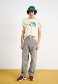 Lys gul bomulds t-shirt med blåt og sort logo, grå corduroy bukser og brune athletic sko. Grøn kasket med broderet detalje.