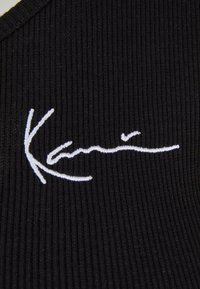 Svart ribbestrikket stoff med en hvit brodert signatur-logo i stil. Teksturen er strukturert og tettsittende, med en subtil glans.