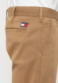 Χακί παντελόνια φτιαγμένα από ανθεκτικό ύφασμα, με λογότυπο Tommy Jeans σε κόκκινο, άσπρο και μπλε στο πίσω τσέπη. Απλός σχεδιασμός, ίσια γραμμή.