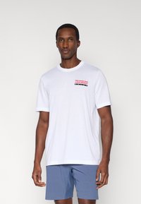 Hvid bomulds-T-shirt med korte ærmer, rund hals, med sort og rød tekst på brystet. Båret med blå shorts.