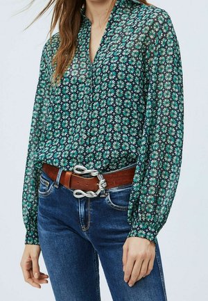 Eine Frau trägt eine türkisfarbene, blumengemusterte Bluse mit Knöpfen, die in eine blaue Jeans gesteckt ist. Um ihre Taille trägt sie einen braunen Ledergürtel mit einer silbernen, gedrehten Schnalle.