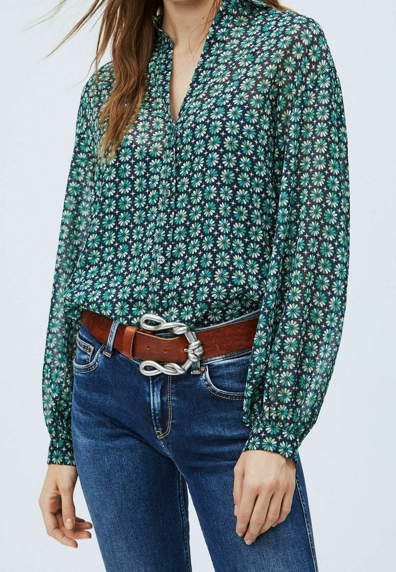 Kobieta w turkusowej, kwiecistej bluzce zapinanej na guziki, włożonej w niebieskie dżinsy, z brązowym skórzanym paskiem z srebrną, skręconą klamrą.