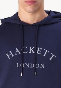 Námořnická modrá mikina s kapucí a stahovací šňůrkou, s velkým světle šedým logem "HACKETT LONDON" na hrudi. Vyrobeno z měkkého materiálu.