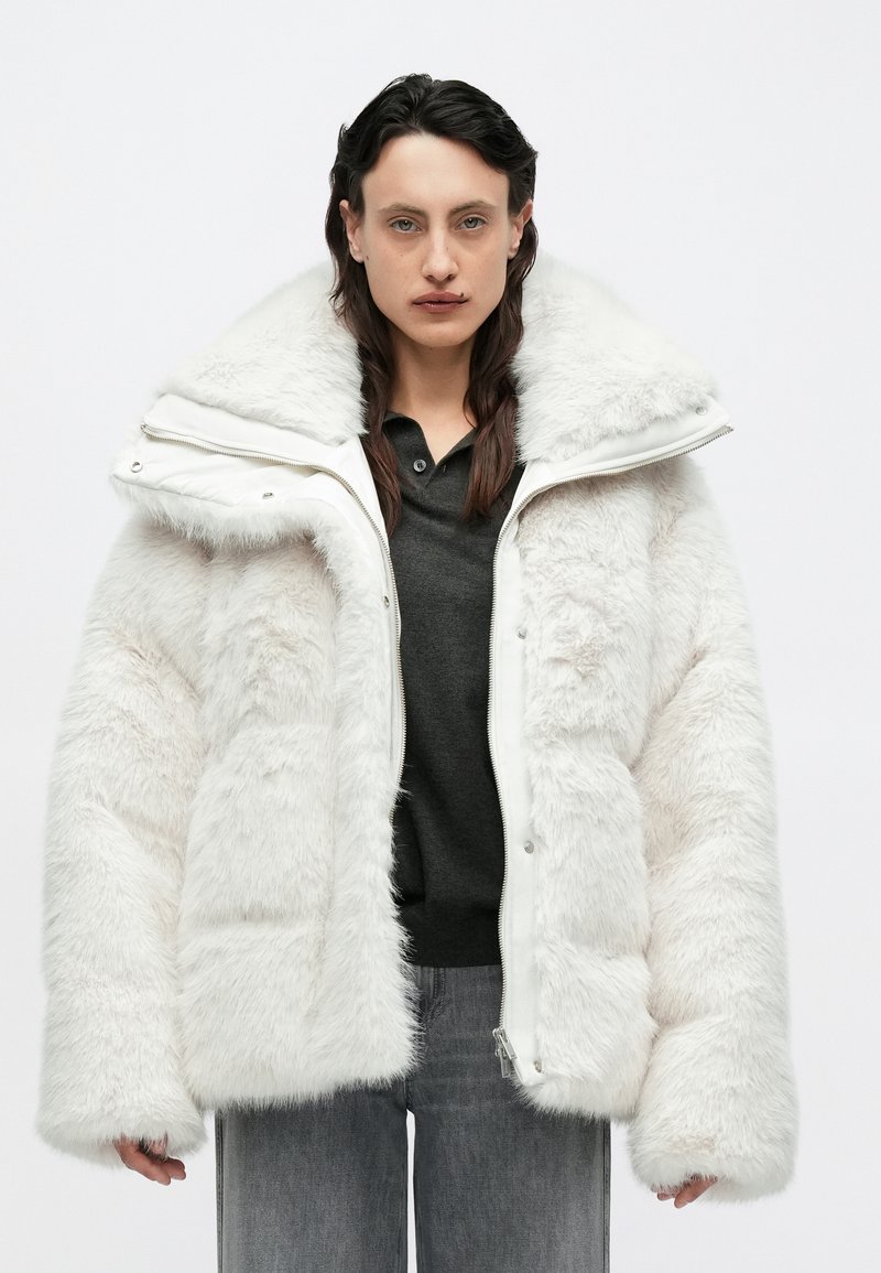Fehér műszőrme steppelt dzseki nagy kapucnival, cipzáras záródással és gombdíszítéssel. Texturált, bolyhos külsővel és széles ujjakkal.