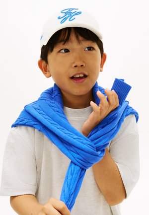 Jaunas berniukas, dėvintis baltą kepurę ir šviesiai pilką marškinėlį, taiso ryškiai mėlyną megztinį su pynuotais raštais, permestą per pečius.