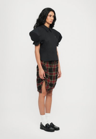 Svart blus med puffärmar i kombination med en knälång rutig kjol med röda och gröna detaljer. Modellen bär svarta loafers och vita strumpor.