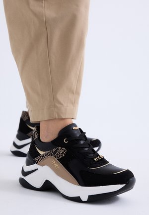 Zapatillas negras y beige con detalle de estampado animal, usadas con pantalones marrón claro sobre un fondo blanco.