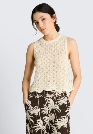 Jeune femme aux cheveux foncés attachés en chignon, portant un top en maille crème sans manches et un pantalon noir avec des motifs de palmiers blancs, les mains dans les poches.