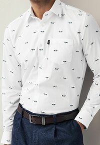 Camisa blanca de manga larga con un patrón repetido de libélulas en azul marino y verde azulado. Presenta un bolsillo en el pecho y un cuello estándar con botones.