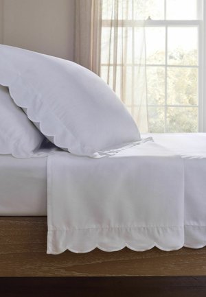 Weißes Bett mit Kissenbezügen und Laken mit gewelltem Rand neben einem Fenster mit transparenten Vorhängen, die natürliches Licht hereinlassen.