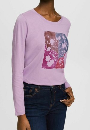 Personne portant une chemise à manches longues violet clair avec un motif floral rose, violet et bleu et un jean bleu foncé.