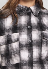 Svart och vit rutigt skjorta i mjukt tyg, med knäppning fram, krage och två bröstfickor med svarta knappar.