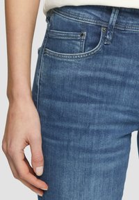 Niebieskie dżinsy o gładkiej fakturze, średnim stanie, z pięcioma kieszeniami i zapięciem na guzik; widoczne szwy i delikatne przetarcia.