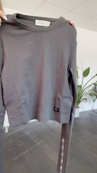 Gray ribbed long-sleeve shirt with a round neck and side slit. Features a small black CK label at the bottom. Smooth texture.