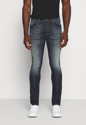 Mann, der ausgewaschene dunkelblaue Slim-Fit-Jeans, ein schwarzes T-Shirt und weiße Turnschuhe trägt, steht vor einem schlichten hellen Hintergrund.