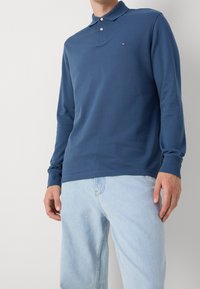 Mężczyzna w granatowej koszulce polo z długim rękawem, w jasnoniebieskich dżinsach, stojący na jednolitym jasnoszarym tle.