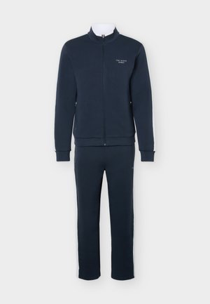 Marineblå sportjakke med glidelås og matchende bukser med hvite side-striper og "Ted Baker Sport" logo på jakken bryst.