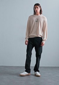 Una persona joven con el cabello largo y oscuro está de pie contra una pared simple, vistiendo un suéter beige, pantalones negros y zapatillas blancas.