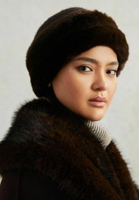 Bruine bonten hoed met een afgerond ontwerp, gedragen met een donkere bontkraag en een geribbelde coltrui. De gladde textuur benadrukt luxe en warmte.