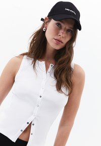 Top sin mangas de canalé blanco con cierres de botón plateados en el frente, combinado con una gorra negra que dice "hey, soy yo."