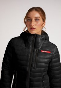 Giacca a vento nera con zip frontale, cappuccio e texture trapuntata verticale. Presenta un'etichetta del marchio rossa sul lato sinistro del petto e tasche con zip.