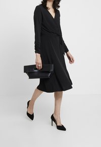 Svart omslagsklänning med långa ärmar, knytmidja och en svängig kjol; kombinerad med en svart clutchny purse och svarta högklackade skor.
