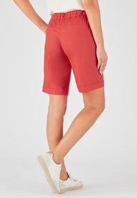 Shorts rojos hasta la rodilla con una textura suave, cinturilla elástica y puños doblados. Presenta dos bolsillos traseros. Combinados con zapatillas blancas.