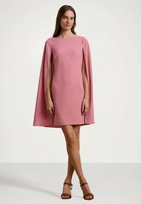 Mulher em pé vestindo um vestido de capa rosa até ao joelho com aberturas nos braços e sandálias de tiras no tornozelo castanhas, contra um fundo simples.