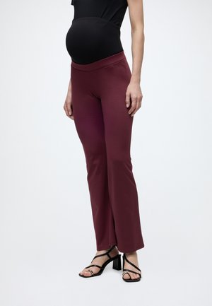 Personne enceinte portant un haut noir, un pantalon de maternité bordeaux et des sandales à talons noires, debout devant un fond clair uni.
