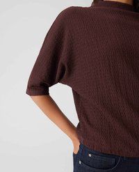Person, die ein strukturiertes, dunkelbraunes, kurzärmliges Oberteil mit umgeschlagenen Ärmeln und dunkelblauen Jeans trägt, mit einer Hand in der Tasche.