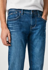 Jeans de mezclilla azul con un corte slim, que presentan un diseño clásico de cinco bolsillos, costuras amarillas en contraste y una pequeña etiqueta de la marca en el bolsillo trasero.