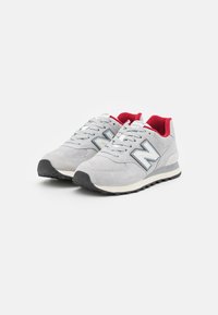 Pár šedých tenisek New Balance s černými podrážkami a červeným vnitřkem, vystavených na bílém pozadí.