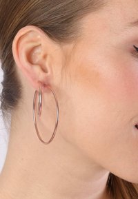 Roségoldene Creolen, die an einem Ohr präsentiert werden. Die Ohrringe verfügen über zwei miteinander verbundene Kreise mit einer glatten, glänzenden Metalloberfläche.