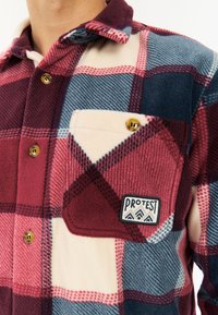 Close-up van een persoon die een fleece shirt met een rood, blauw en crèmekleurig ruitpatroon draagt, met bruine knopen en een 'Protest'-patch op de borstzak.