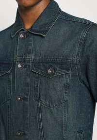 Dunkelblaues Denim-Jackett mit klassischem Kragen, Brusttaschen und silbernen Knöpfen. Texturiertes Material mit sichtbaren Nähten.