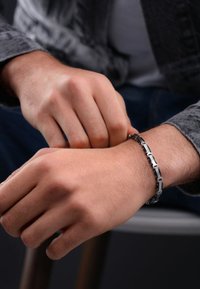 Bracciale in metallo con maglie rettangolari, sezioni alternate in argento e nero. Texture liscia, indossato attorno a un polso, mostrato in un contesto informale.