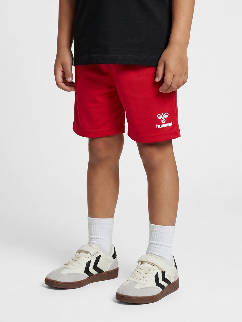Kind trägt rote hummel Shorts, weiße Socken und weiße Sneakers mit schwarzen Streifen auf einfachem Hintergrund.