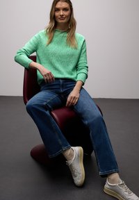 Femme assise sur une chaise moderne bordeaux, portant un pull vert, un jean bleu, des chaussettes beige et des baskets blanches, sur un fond gris.