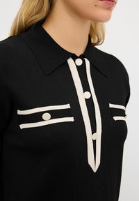 Maglietta polo nera in maglia con design a strisce verticali bianche, dotata di due tasche sul petto, bottoni e colletto classico.