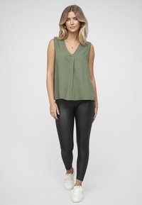 Olivengrønn ermeløs topp med V-hals, kombinert med skinnende svarte leggings og hvite joggesko. Enkle og rene linjer gjennomgående.