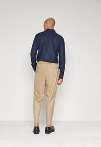 Marinblå långärmad skjorta med knappade manschetter som är ihopparad med ljusbeige elastiska byxor med midjeresår och svarta skor, sedd bakifrån.