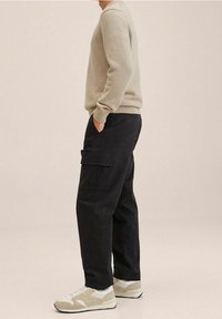Beige gestrickter Pullover, schwarze Cargo-Hosen mit Seitentaschen und beige Sneaker mit weißen Akzenten. Strukturiertes Material, lockere Passform.